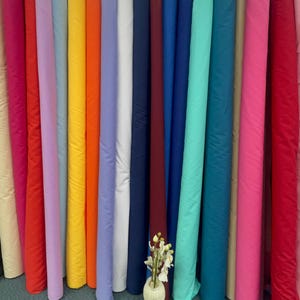 May include: A display of fabric rolls in a range of colours, such as pink, red, orange, yellow, blue, and green. A small vase with flowers is placed in front of the rolls. The fabric is tightly rolled and appears to be a solid colour.