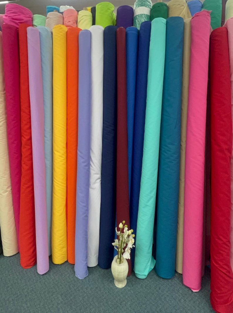 May include: A display of fabric rolls in a range of colours, such as pink, red, orange, yellow, blue, and green. A small vase with flowers is placed in front of the rolls. The fabric is tightly rolled and appears to be a solid colour.