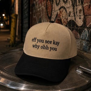 May include: A two-tone baseball cap featuring a tan crown and a black brim. The front of the crown displays black embroidered text that reads, "eff you see kay why ohh you" arranged in two lines. The cap has a structured front panel and a button.