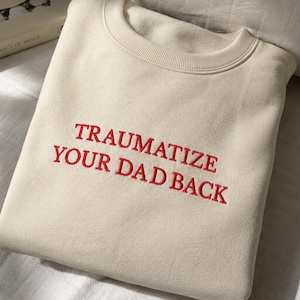 May include: A cream-colored crewneck sweatshirt featuring the text "TRAUMATIZE YOUR DAD BACK" embroidered in red capital letters across the chest. The garment has a ribbed neckline and long sleeves, presented in a folded, flat-lay orientation.