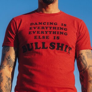 May include: A red short-sleeved t-shirt featuring black text printed on the chest that reads: DANCING IS EVERYTHING EVERYTHING ELSE IS BULLSHIT. The shirt has a crew neckline and a standard fit, displayed against a clear blue sky background.