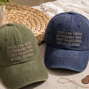 May include: An olive green baseball cap and a navy blue baseball cap, both featuring embroidered text that reads: YOUR TWO CENTS IS PROBABLY WHY THEY'RE GETTING RID OF PENNIES. Each cap has a structured crown, a curved brim, and matching eyelets.