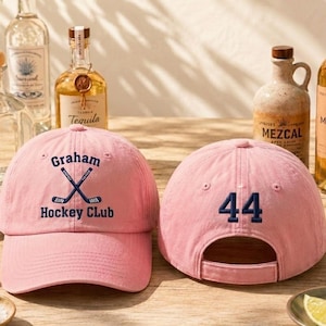May include: Two pink baseball caps, one with "Graham Hockey Club" and crossed hockey sticks, the other with the number "44" in navy blue. Tequila and mezcal bottles are in the background, along with limes and salt.