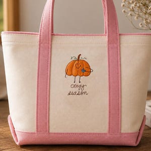 May include: A cream-colored canvas tote bag with pink trim and handles. The front features an embroidered orange pumpkin character holding a blue mug, with the text "cozy season" stitched in dark brown thread below the pumpkin graphic.