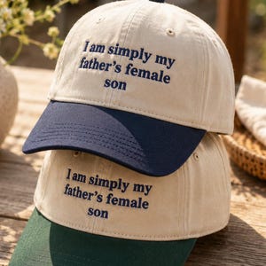 May include: Two baseball caps stacked on top of each other. The caps are beige with navy blue and green brims. Each cap has the embroidered text: "I am simply my father's female son."