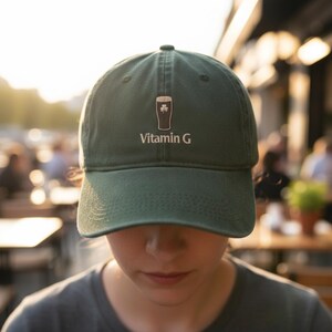 May include: A dark green baseball cap with a curved brim. The cap features the text "Vitamin G" in white, along with a graphic of a pint of beer with a shamrock. The cap is made of a soft material.