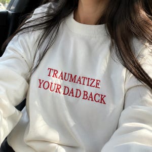 May include: A white long-sleeve crewneck sweatshirt featuring the text "TRAUMATIZE YOUR DAD BACK" embroidered in red capital letters across the chest. The garment has a ribbed neckline and long sleeves, providing a simple and clean aesthetic.