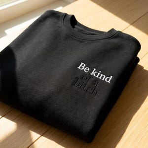May include: Black crewneck sweatshirt with the embroidered phrase "Be kind of a bitch" in white. The sweatshirt is folded neatly on a wooden surface, with natural light illuminating the fabric.