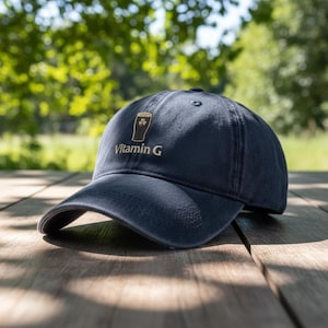 May include: Navy blue baseball cap with a curved brim. The cap features an embroidered design of a beer glass with a shamrock and the text "Vitamin G" in white. The cap is sitting on a wooden surface.
