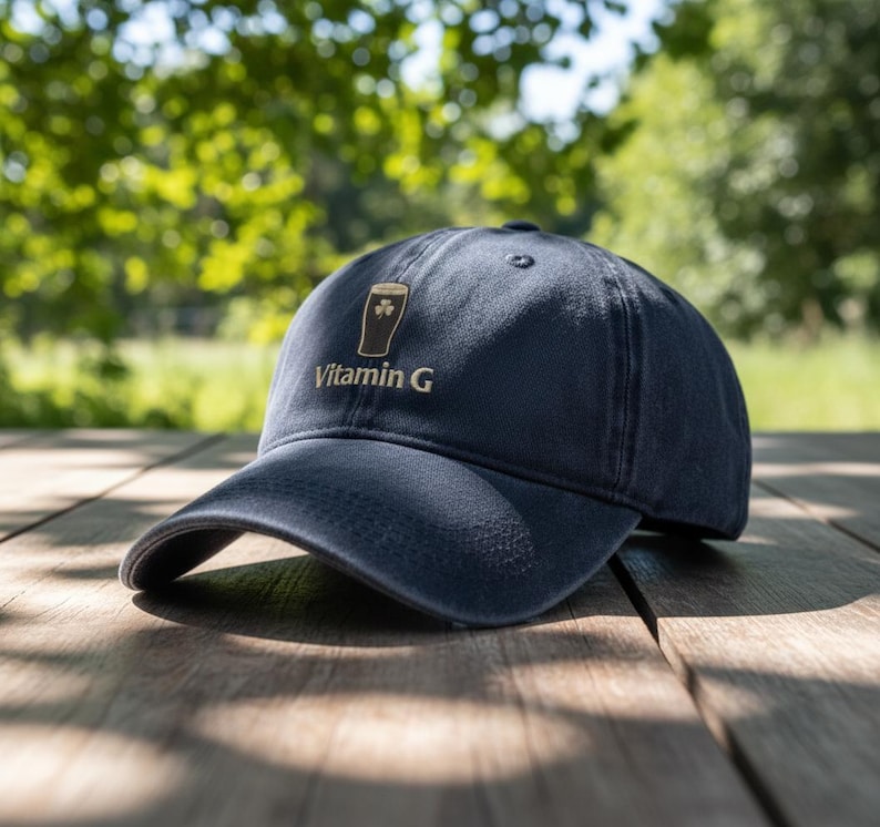 May include: Navy blue baseball cap with a curved brim. The cap features an embroidered design of a beer glass with a shamrock and the text "Vitamin G" in white. The cap is sitting on a wooden surface.