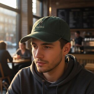 May include: A dark green baseball cap with the text "Vitamin G" in white. The cap has a curved brim and a small graphic. The person in the image is wearing a gray hoodie.