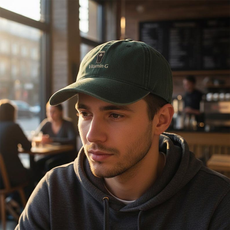 May include: A dark green baseball cap with the text "Vitamin G" in white. The cap has a curved brim and a small graphic. The person in the image is wearing a gray hoodie.