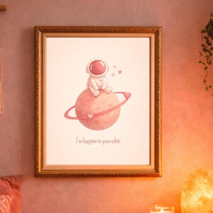 Puede incluir: Una impresión enmarcada que presenta a un astronauta sentado en un planeta rosa con un anillo en forma de corazón. La impresión tiene el texto "I'm happier in your orbit" en un marco dorado. La obra de arte se exhibe en una pared rosa.