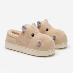 May include: A pair of beige hippo-shaped slippers. The slippers have a plush texture, with grey accents for the ears and nostrils, and black eyes. The soles are a light cream colour.