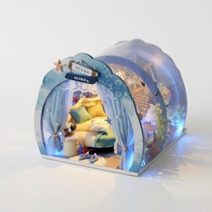 Może przedstawiać: Miniaturowy domek dla lalek o tematyce oceanicznej z zakrzywionym, przezroczystym dachem. Niebiesko-biała konstrukcja przedstawia scenę sypialni z łóżkiem, poduszkami i zasłonami. Znak głosi "Ocean Homes". Oświetlenie LED oświetla wnętrze.
