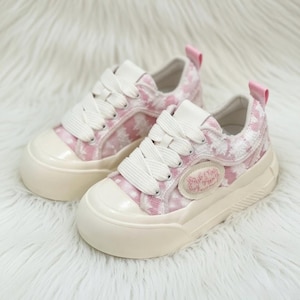 Może przedstawiać: Para różowo-białych płóciennych tenisówek z białymi sznurowadłami i kremową podeszwą. Buty mają różową pętelkę i małą naszywkę z tekstem. Na białej, puszystej powierzchni.