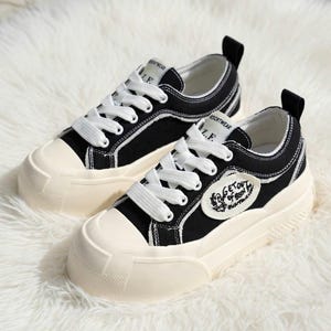 以下が含まれることがあります： 白の靴紐と厚いオフホワイトのソールが付いた、黒色のキャンバススニーカーのペア。 スニーカーには白いトリムと「GET OUT OF BED」と書かれたパッチが付いています。 白くふわふわした表面にあります。