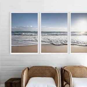 Puede incluir: Tres fotografías enmarcadas de una escena de playa con olas blancas y espumosas rompiendo en la orilla. Las fotos están colgadas en una pared blanca con dos sillas de mimbre delante de ellas.
