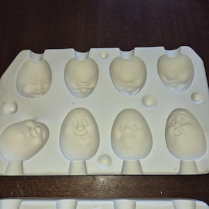 May include: A white silicone mould with eight egg-shaped cavities, each featuring a different facial expression. The mould is designed for crafting or baking, with a variety of whimsical faces. The mould is made of a flexible, food-safe material.