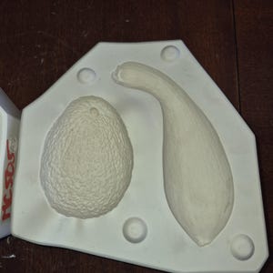 May include: A white, geometric mould featuring two cavities: one shaped like a textured avocado and the other like a curved squash. The mould has several circular indentations and is set against a dark wooden surface. The brand name "rcsos" is visible on the side.