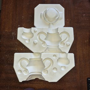 May include: White ceramic moulds for creating decorative objects. The moulds feature designs of vases, handles, and circular accents. The moulds are arranged on a dark brown wooden surface.