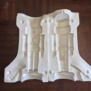 May include: Two-part white plaster mould for a nutcracker figurine. The mould features detailed relief impressions of a traditional nutcracker soldier, including uniform details and a sword. The mould is designed for creating decorative holiday items.
