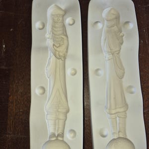 May include: Two white ceramic moulds, each depicting a detailed Santa Claus figure. The Santa figures are shown in a classic pose, holding a small object. The moulds are set against a neutral background, highlighting the intricate design.