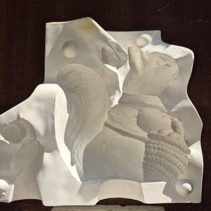 May include: A white sculpture of a squirrel with a basket of fruit, set against a backdrop of stylized cheese. The squirrel has a fluffy tail and a collar. The sculpture is mounted on a dark brown surface.
