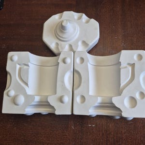 May include: A collection of white ceramic mug moulds. The set includes two halves for the mug body, a base, and a lid. The pieces are arranged on a dark brown wooden surface. The moulds are for creating a mug.