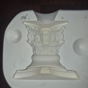Peut inclure: Un moule en plâtre blanc représentant une colonne classique. La colonne présente des détails complexes, notamment des volutes ornées et une base décorative. Le moule est inséré dans une forme ovale plus grande, avec un sommet incurvé et deux creux circulaires.