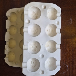 May include: Two off-white, plastic moulds for making round objects. Each mould has a series of circular indentations. The moulds are on a dark brown wooden surface. The indentations have small, raised designs.