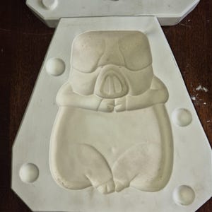May include: An off-white, two-part mould featuring a stylized Stormtrooper design. The mould is triangular with rounded edges and circular indentations. The Stormtrooper's helmet and body are clearly defined in the mould.