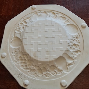 May include: Off-white, octagonal ceramic trivet with a woven basket design in the centre. The border features embossed fruit, including grapes, pears, and strawberries. Small, rounded feet are placed on the corners.