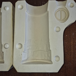 May include: Off-white plaster mould with a lighthouse design. The mould is in three parts, with a cylindrical shape and a detailed relief of a building with a tower. The mould has a smooth surface with circular indentations.