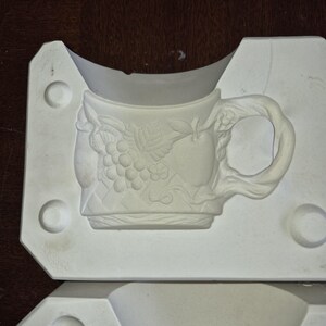 May include: A white ceramic mould for a decorative mug. The mug design features embossed grapes, an apple, and floral accents. The handle has a branch-like appearance. The mould is set in a rectangular frame with circular indentations.