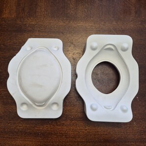 May include: Two white, oval-shaped moulds with rounded edges and small, circular details. One mould is solid, while the other has a central opening. The moulds are likely for crafting or casting small objects.