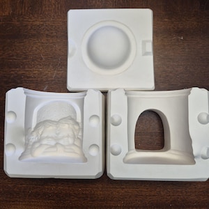 Peut inclure: Trois moules en céramique blanche pour l'artisanat. L'un des moules présente une indentation circulaire, tandis que les deux autres présentent des motifs en relief détaillés de chérubins et une fenêtre arquée. Les moules sont disposés sur une surface en bois.