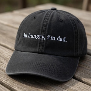 May include: A black baseball cap featuring white embroidered text on the front that reads, hi hungry, i'm dad. The cap has a curved brim, a structured crown with stitched eyelets, and a button at the top center of the fabric panels.