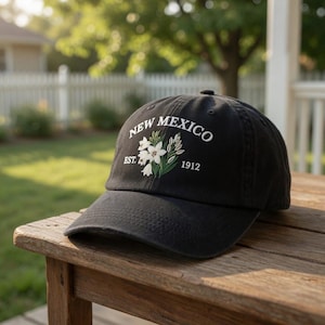 May include: Black baseball cap with the text "NEW MEXICO" in white, along with an embroidered floral design and the year "1912". The cap is sitting on a wooden surface.
