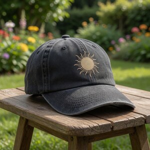 May include: A black baseball cap with a gold sun embroidered on the front. The cap is sitting on a wooden stool in a garden setting. The cap has a curved brim and a fabric strap closure.