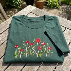 May include: A dark forest green short-sleeve t-shirt featuring an embroidered design of various red mushrooms with white spots, interspersed with green stems, leaves, and small yellow floral accents across the lower front section.