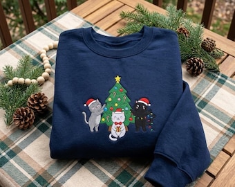 May include: A navy blue crewneck sweatshirt featuring an embroidered graphic of three cats wearing Santa hats around a decorated Christmas tree. One gray cat, one white cat holding a gift, and one black cat tangled in colorful string lights.