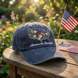 May include: A charcoal grey baseball cap featuring an embroidered outline of the United States filled with colorful wildflowers and the white cursive text America the Beautiful. The cap has a curved brim and a structured crown with eyelets.