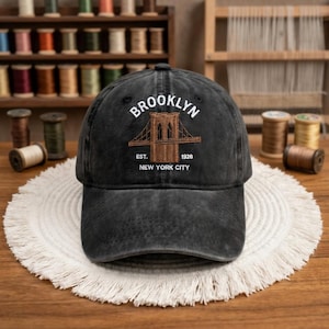 May include: Black baseball cap with the word "BROOKLYN" embroidered in white above a brown illustration of the Brooklyn Bridge. The text "EST. 1928 NEW YORK CITY" is below the bridge. The cap is on a white, fringed mat.