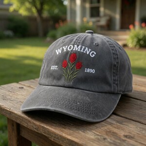 May include: A gray baseball cap with the word "WYOMING" embroidered in white, along with the text "EST. 1890". The cap features a floral design with red flowers and green stems. The hat is sitting on a wooden surface.