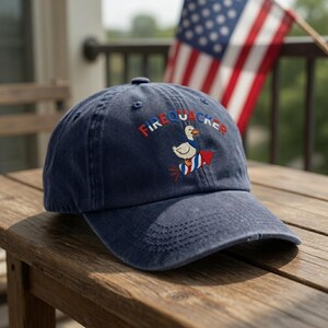 May include: Navy blue baseball cap with the word "FIREQUACKER" embroidered in red, white, and blue. The cap features an embroidered cartoon duck wearing a cape and holding a firework. The cap is sitting on a wooden table with an American flag in the background.