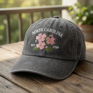 Op de afbeelding: Een donkergrijze baseballcap met de tekst "NORTH CAROLINA" in witte letters geborduurd boven een bloemenontwerp. Het ontwerp toont roze bloemen en groene bladeren. De tekst "EST. 1789" is ook op de cap geborduurd.