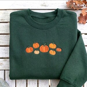 May include: A dark forest green crewneck sweatshirt featuring an embroidered design of seven orange pumpkins of varying sizes arranged in a horizontal cluster across the center of the chest. The pumpkins have small brown stems and ribbed textures.