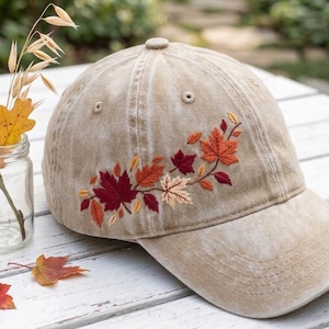 May include: A tan baseball cap featuring embroidered autumn leaves in shades of deep red, burnt orange, and cream. The cap has a distressed, washed-out texture and a curved brim, with the leaf design positioned on the side panel.