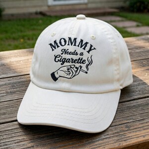 May include: Cream-colored baseball cap with the embroidered text "MOMMY Needs a Cigarette" above a graphic of a hand holding a lit cigarette. The cap has a curved brim and a fabric strap closure.